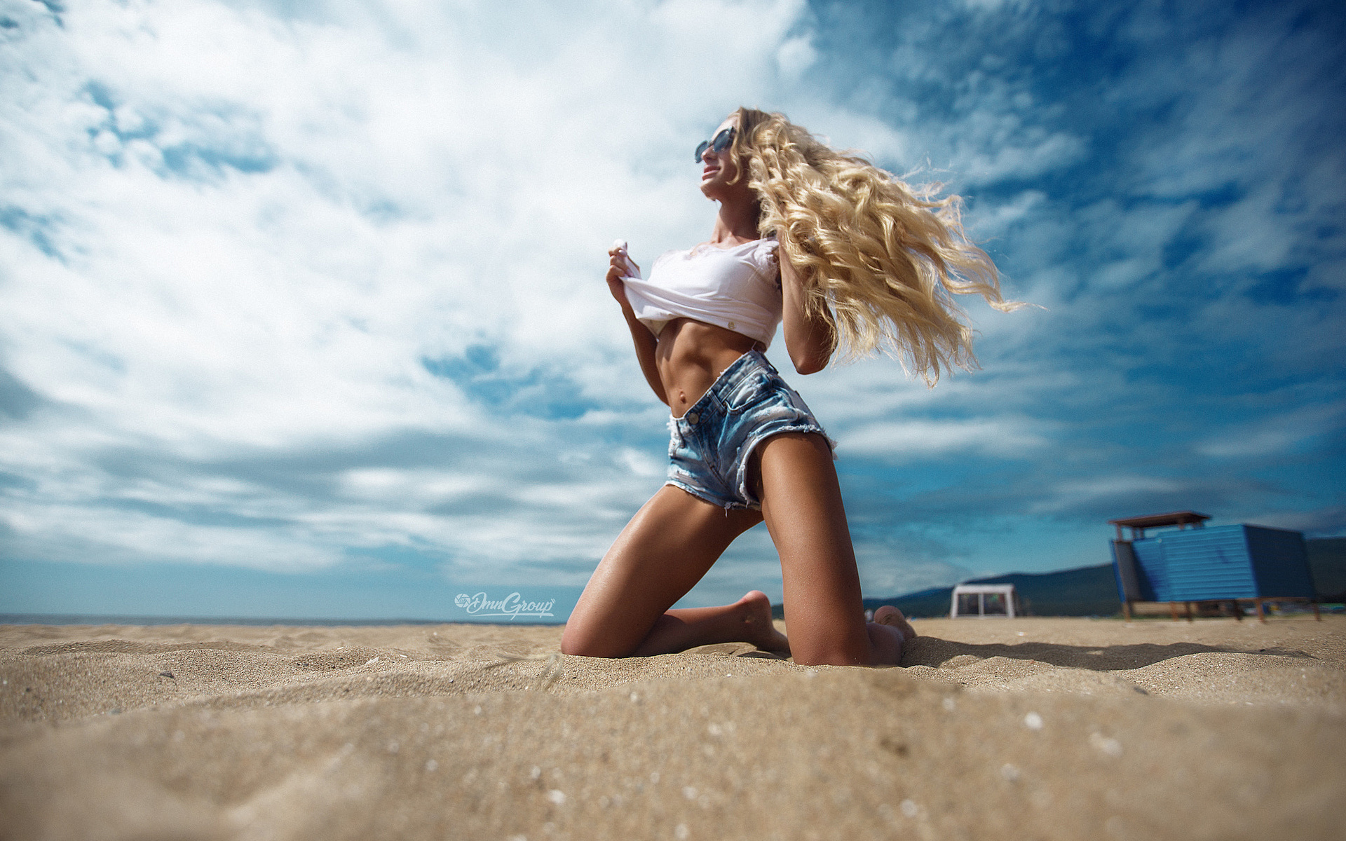women, blonde, belly, sand, t-shirt, tanned, jean shorts, sky, clouds, sunglasses, kneeling