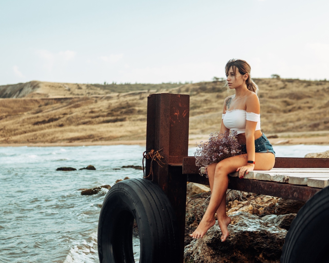 women, brunette, sea, women outdoors, jean shorts, pier, sitting, red nails, tattoo, sky, watch, tires, bare shoulders, looking away